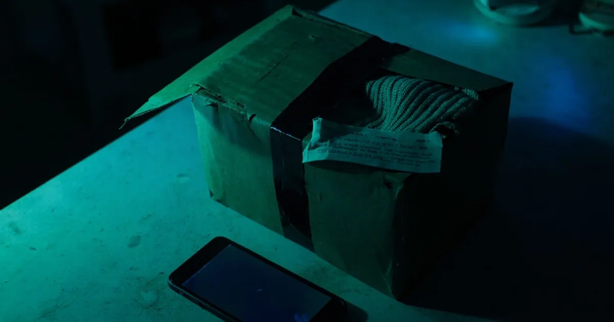 A slightly crushed brown cardboard parcel with a torn corner and packing tape, with a knitted sweater peeking out, lying on a dark concrete table next to a phone with a blurred screen